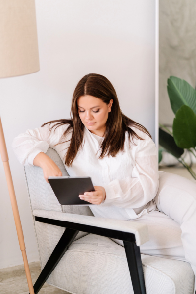Woman with brown hair in a white outfit sits in a chair and looks at a tablet, searching for how to get traffic to your website without social media, next to a floor lamp and a green plant.