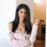 Woman with long dark hair in an off-shoulder light pink top stands indoors, smiling at the camera with her left hand slightly raised—perfect for a conscious copywriter’s profile picture.