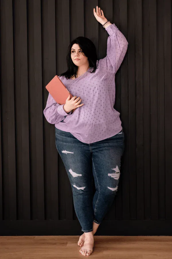 Woman with dark hair wearing a lavender blouse and ripped jeans stands barefoot against a dark wall, holding a closed pink book—capturing the creative spirit behind our b2b copywriting services.