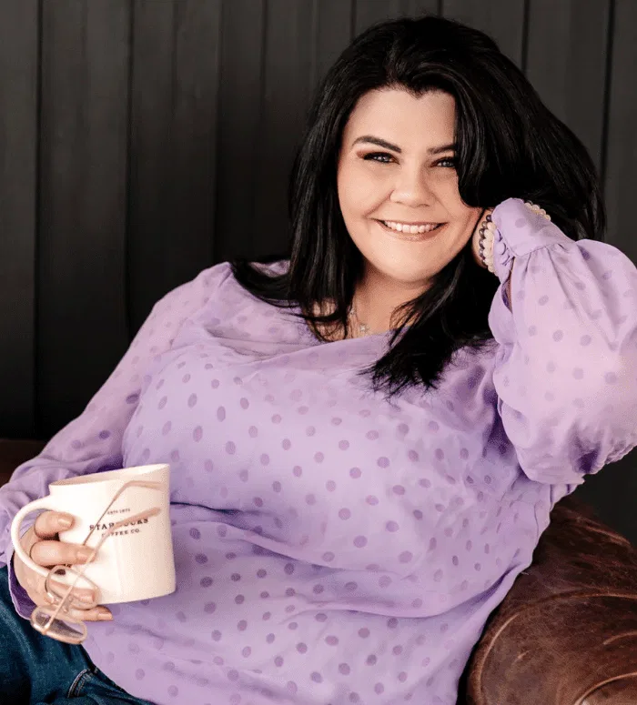 A woman with long dark hair in a sheer lavender polka-dot blouse sits on a couch, holding a white mug and smiling at the camera—radiating the warmth and creativity of a conscious copywriter.