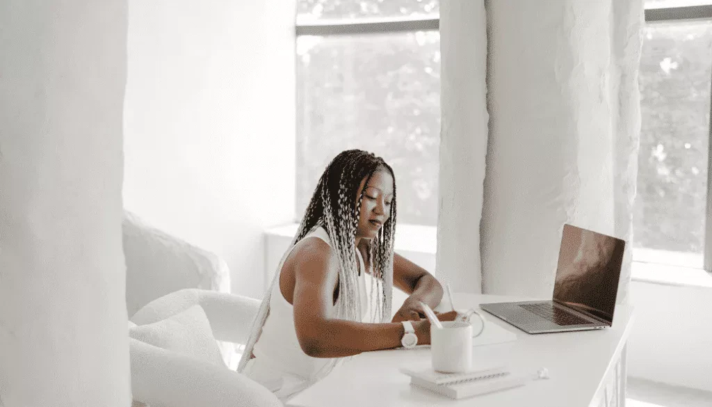 A woman sits at a white desk writing in a notebook, with a laptop, a spiral notepad, and a mug beside her in a bright, minimalist room.