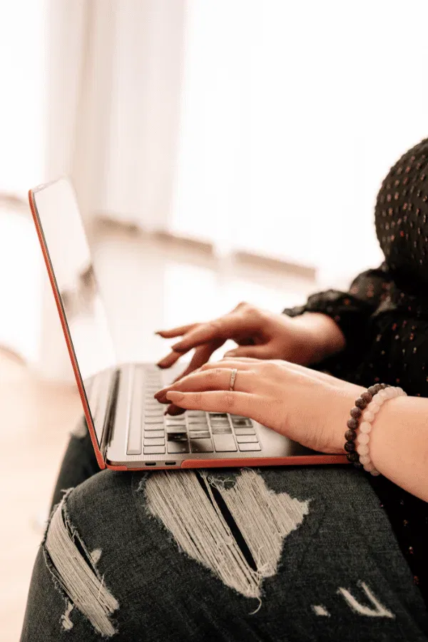 A creative copywriter in a black top and ripped jeans types on a laptop placed on their lap.