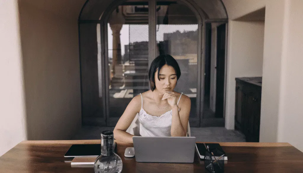 A woman sits at a wooden table working on a laptop, with a notebook, tablet, and water bottle nearby in a bright, airy room.