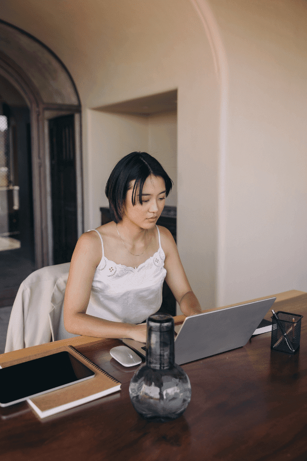 A woman sits at a wooden desk working on a laptop, with a notebook, tablet, and glass vase also on the desk.