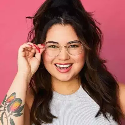 A woman with long dark hair and glasses smiles at the camera, holding her glasses with one hand. Her floral tattoo hints at a creative copywriters touch, while her light gray sleeveless top adds to her relaxed, approachable vibe.