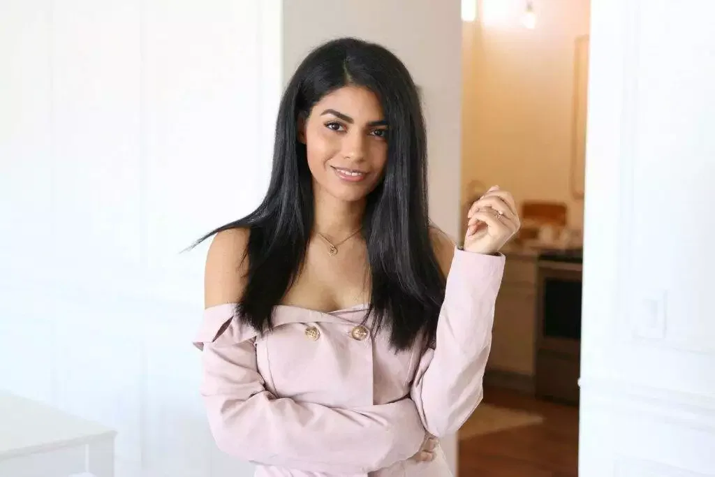 Woman with long dark hair in an off-shoulder light pink top stands indoors, smiling at the camera with her left hand slightly raised—perfect for a conscious copywriter’s profile picture.