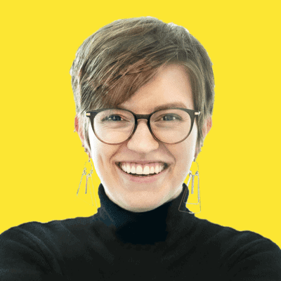 Person with short brown hair, glasses, and earrings smiling, wearing a black turtleneck, in front of a bright yellow background—an approachable conscious copywriter ready to bring ethical copywriting to your brand.
