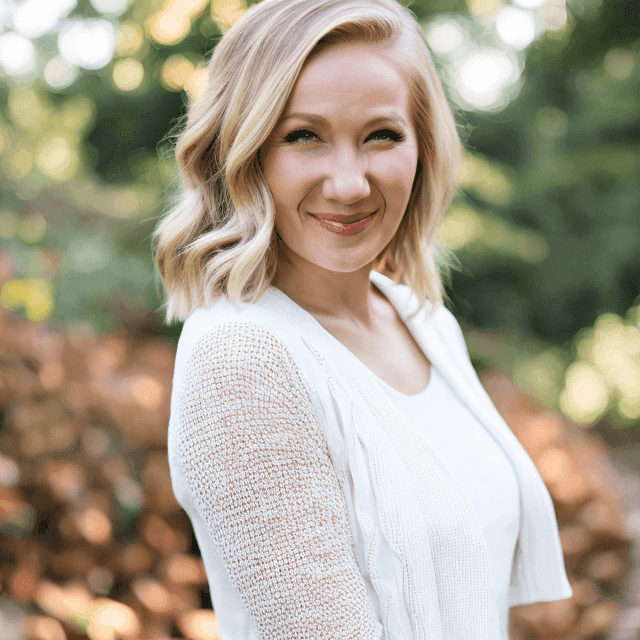 A creative copywriter with blonde, shoulder-length hair and a white sweater stands outdoors, smiling at the camera. Trees and foliage are blurred in the background.