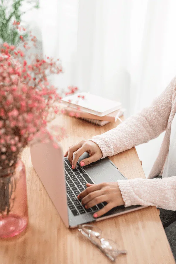 A conscious copywriter types on a laptop at a wooden desk, surrounded by a pink vase of flowers, notebooks, and glasses.