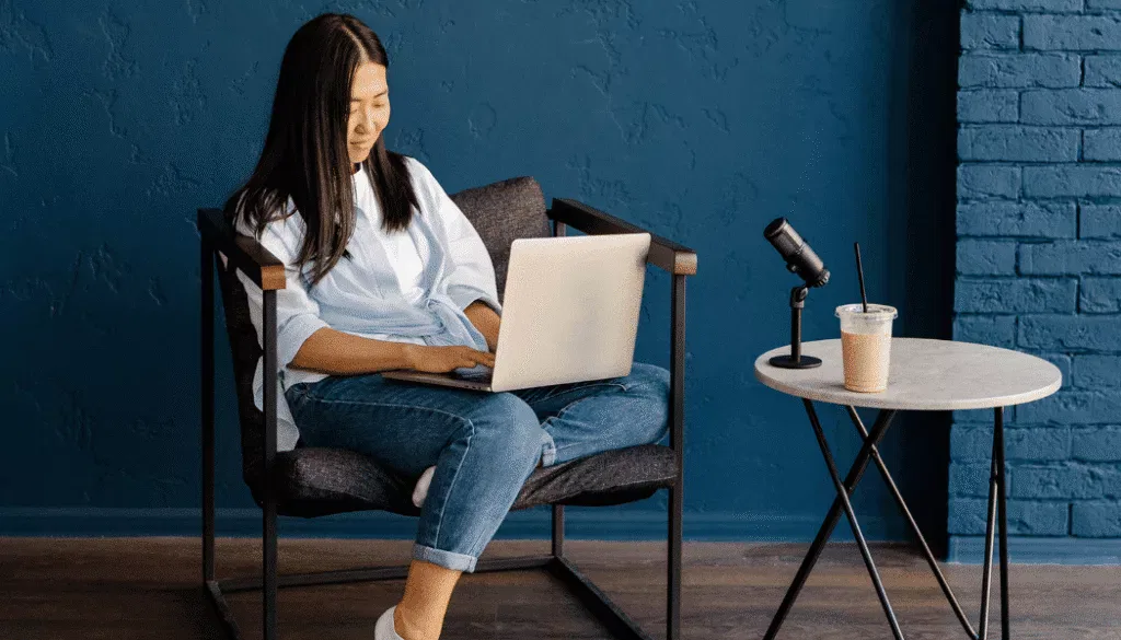 A person sits in a chair using a laptop, with a microphone and a cup of iced coffee on a round side table nearby against a blue wall.
