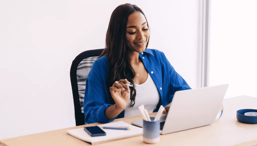 Woman in blue shirt sits at a desk using a laptop, with a notebook, phone, and cup of pens nearby.