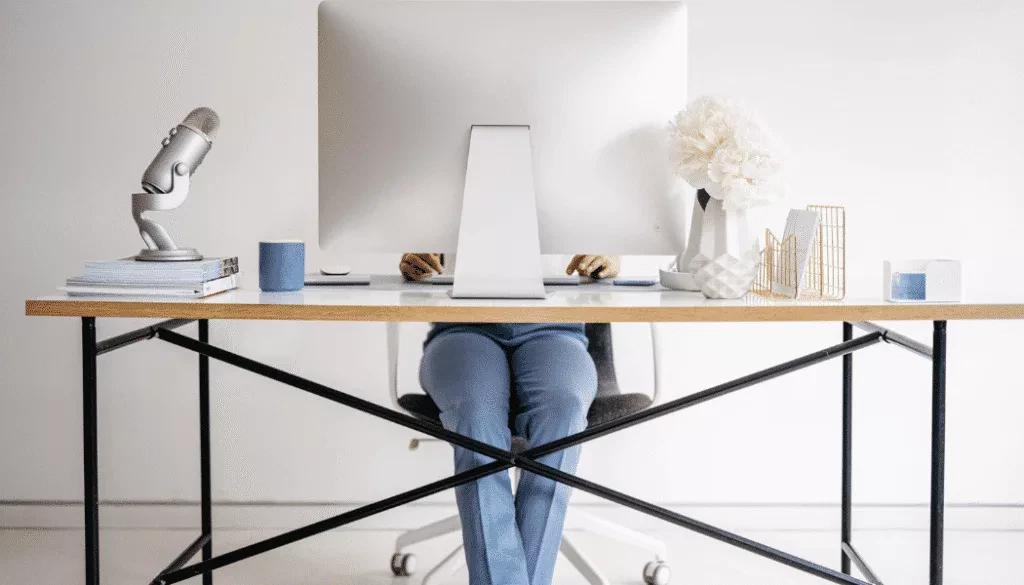 Person sitting at a desk working on a computer, with a microphone, books, mug, decorative flowers, and office supplies on a tidy desktop.