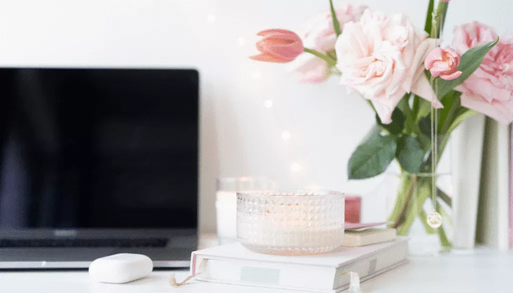 A laptop, a closed notebook, a candle, and a vase of pink flowers sit on a white desk with soft lighting in the background.