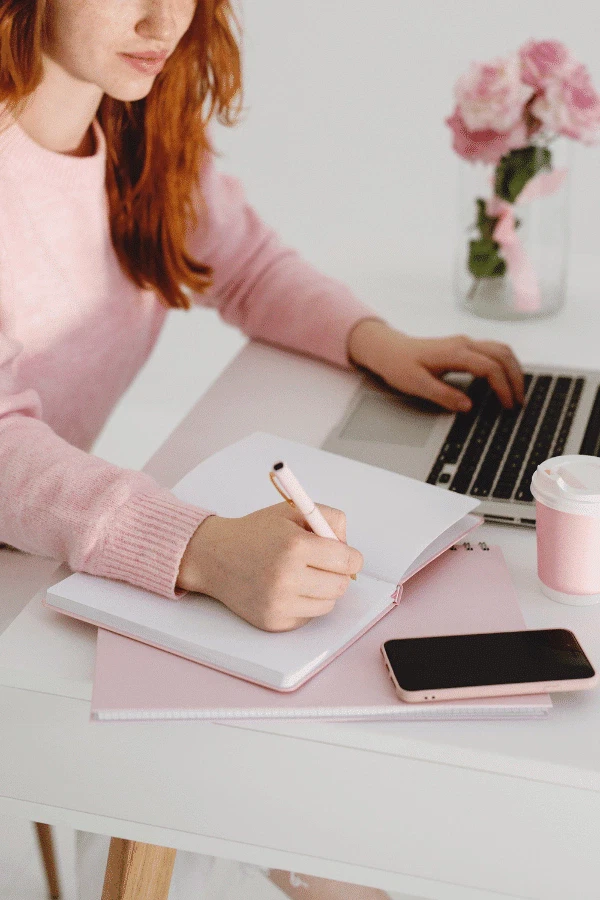 A person in a pink sweater writes in a notebook and uses a laptop at a white desk with a smartphone, coffee cup, and vase of pink flowers.