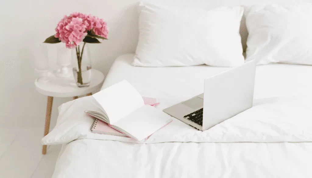 A laptop, an open notebook, and a pen are on a white bed next to a side table with pink flowers in a vase.