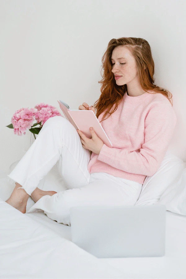 A woman in a pink sweater and white pants sits on a bed with a notebook, next to pink flowers and a laptop.