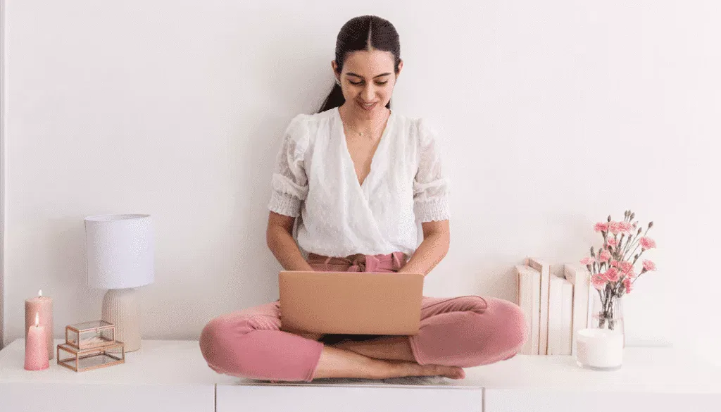 A woman sits cross-legged on a white surface, using a laptop. She is surrounded by candles, a lamp, books, and a vase of pink flowers.