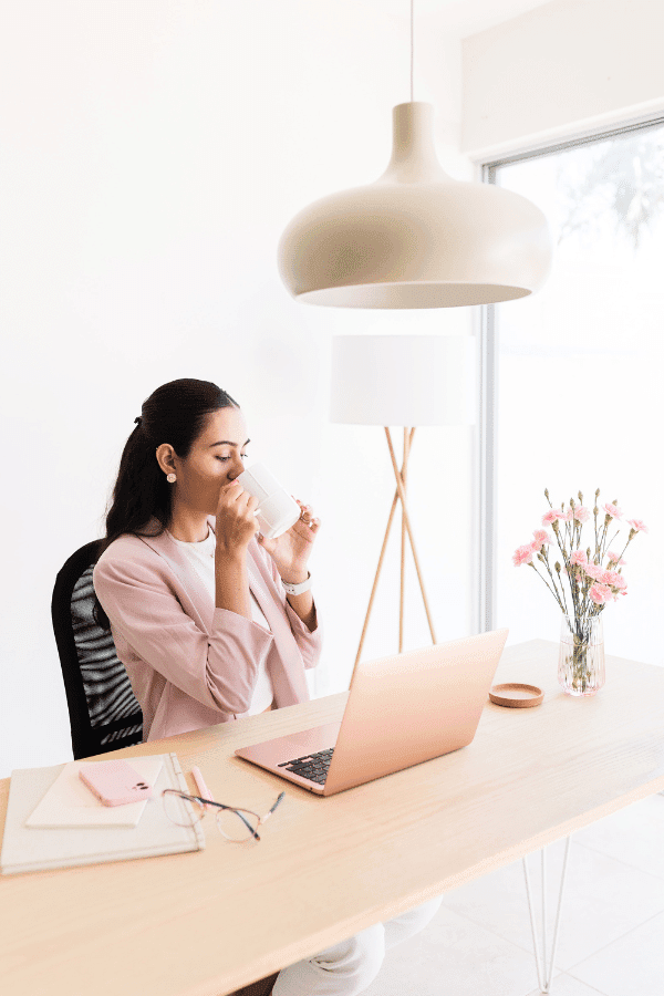 A woman sits at a desk with a pink laptop, drinking from a white mug. The desk also has notebooks, glasses, a pen, and a vase of pink flowers.