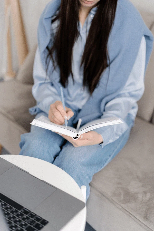 A person sitting on a couch writes in a notebook with a pen, with a laptop open on a nearby table.
