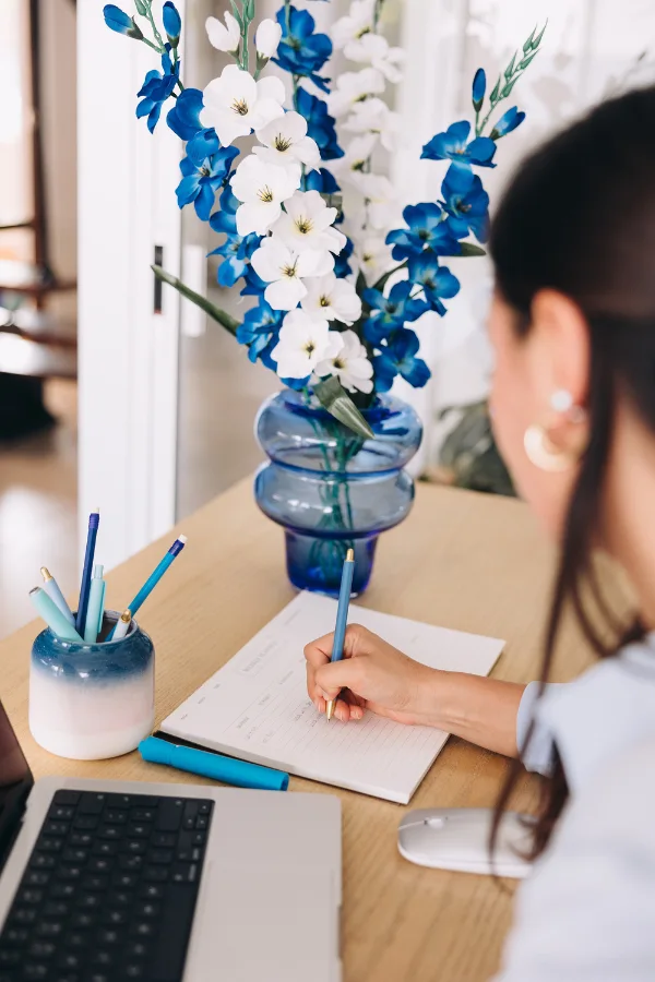 A person writes in a notebook at a desk with a laptop, mouse, pen holder, and a vase of blue and white flowers.