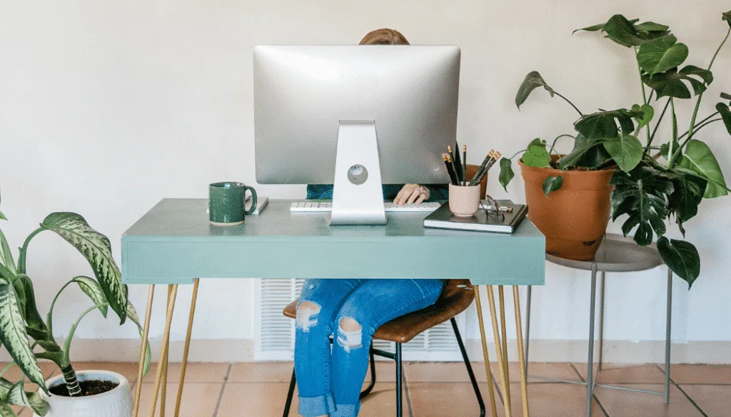 A person sitting at a green desk working on a desktop computer, surrounded by potted plants and office supplies.