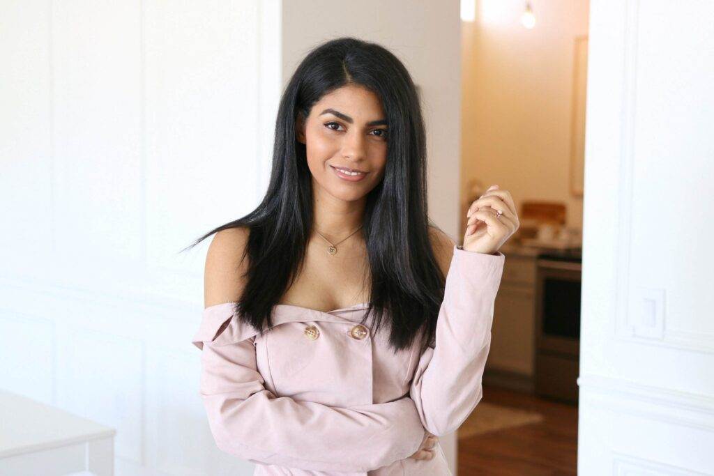 Woman with long dark hair in an off-shoulder light pink top stands indoors, smiling at the camera with her left hand slightly raised—perfect for a conscious copywriter’s profile picture.