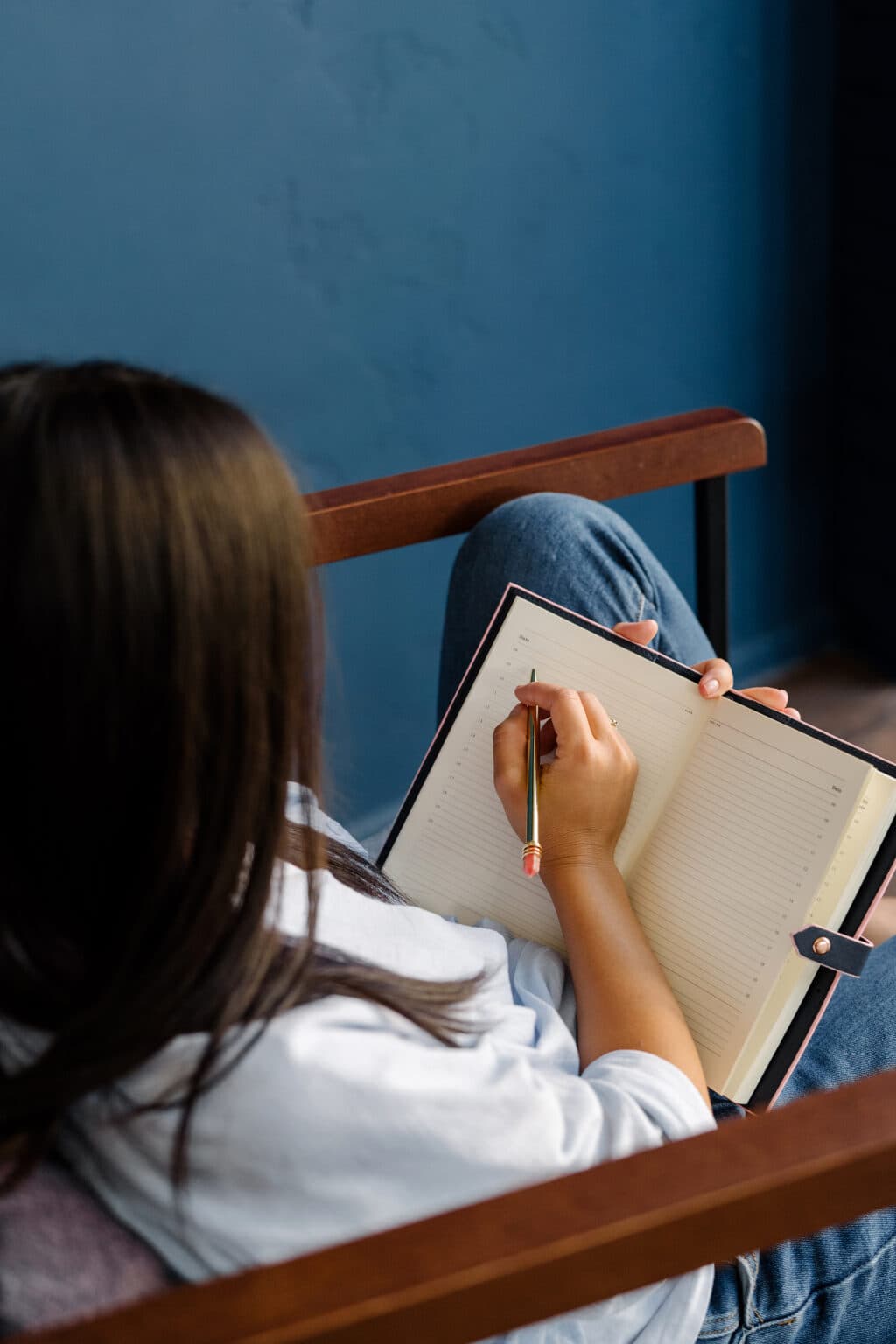 A person is sitting in a chair, writing in a lined notebook with a pencil—perhaps brainstorming ideas for copywriting services. The notebook is open on their lap, and the background wall is blue.