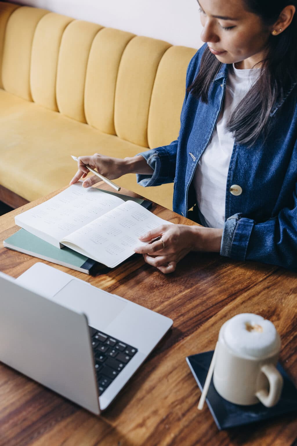 Person sitting at a wooden table with a laptop, planning in a notebook with a pen, next to a cup of coffee topped with foam, thoughtfully considering their next project and exploring professional copywriting services.