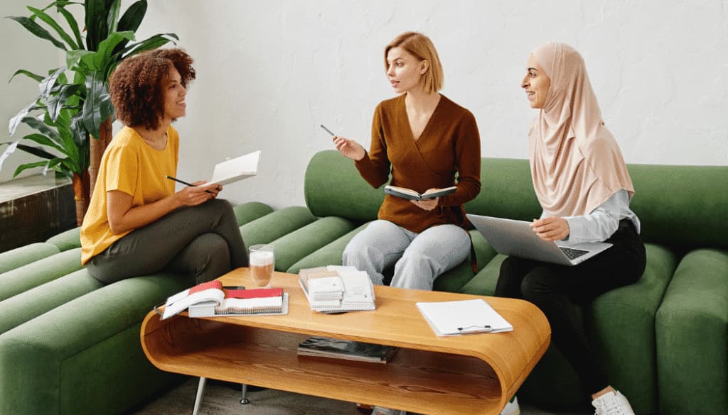 Three women sit on green sofas in an office, talking and taking notes with notebooks and a laptop on a wooden coffee table between them.
