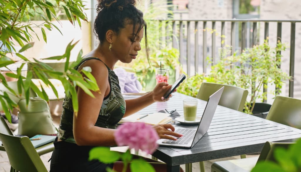 A woman sits at an outdoor table with a laptop and phone, working beside a notebook and a drink, surrounded by greenery—perhaps researching what is website copywriting as she drafts engaging content in the fresh air.
