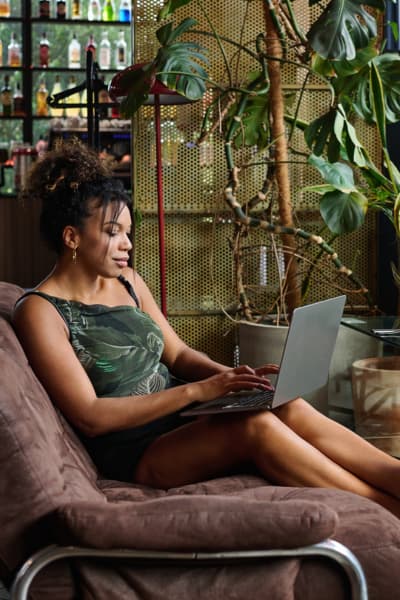 Woman sitting on a brown couch, using a laptop in a modern indoor setting with plants and shelves of bottles in the background, perhaps researching what is website copywriting as she works comfortably from home.