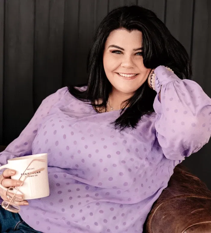 Woman with long dark hair wearing a lavender polka dot blouse sits on a brown chair, smiling, holding a mug and a pair of glasses—her joyful expression says thank you for your purchase.