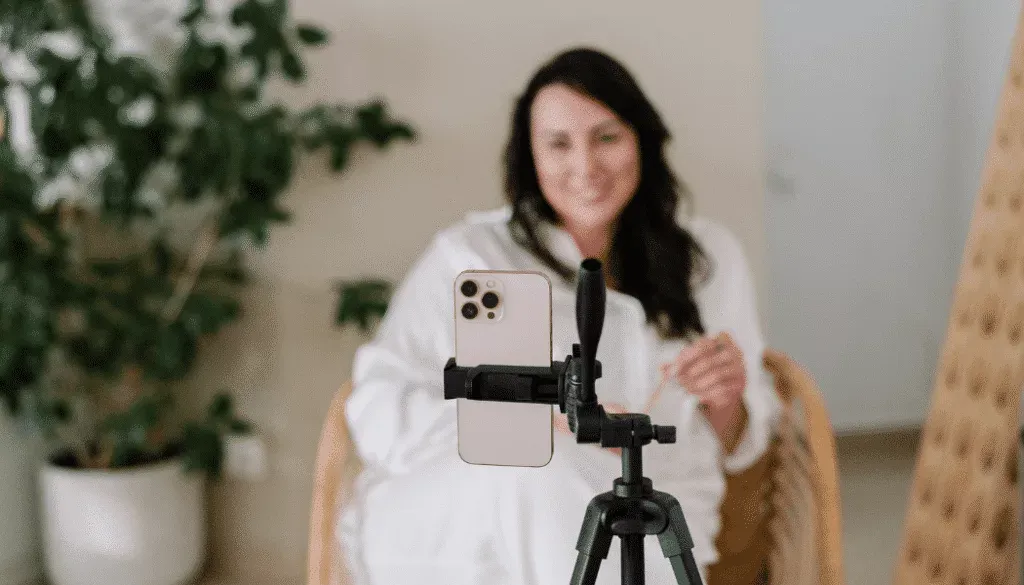 A woman sits in a chair, smiling and talking in front of a smartphone mounted on a tripod, with a potted plant in the background.