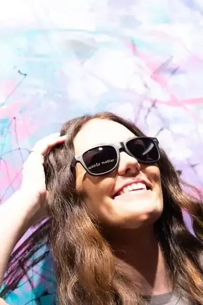 A woman with long brown hair smiles while wearing black sunglasses, standing in front of a colorful, abstract painted wall—radiating the creativity of an ethical copywriter.