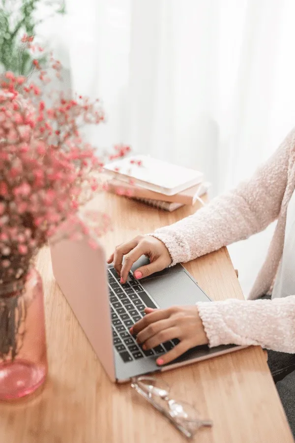 A conscious copywriter types on a laptop at a wooden desk, surrounded by a pink vase of flowers, notebooks, and glasses.