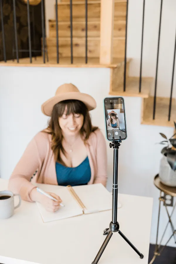 Woman wearing a hat sits at a desk, writing in a notebook, while filming herself with a smartphone on a tripod. A mug and a plant are on the table.