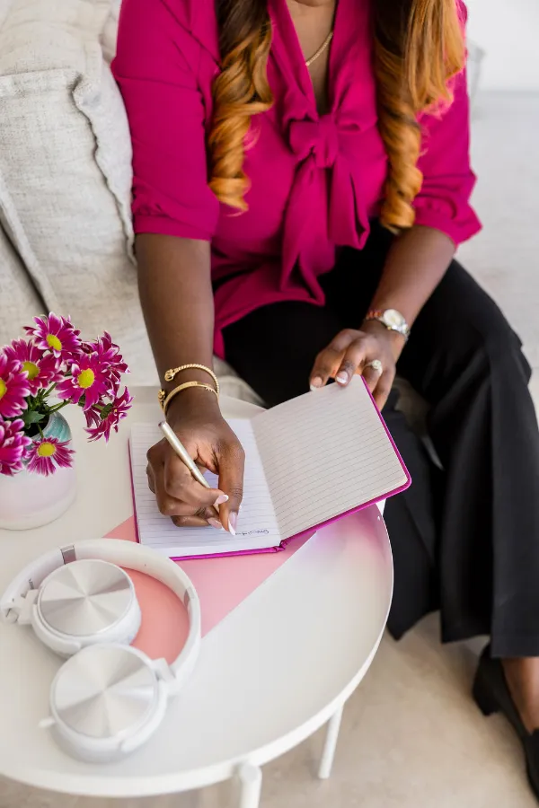 A person in a magenta blouse writes in a notebook while sitting on a couch next to a table with flowers, headphones, and a laptop.