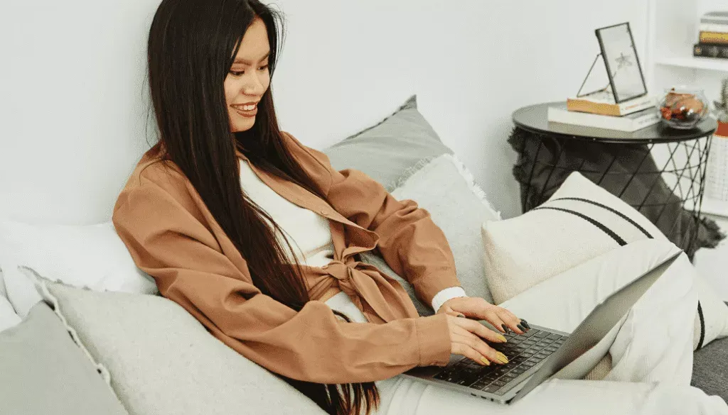 A woman sits on a sofa, smiling and typing on a laptop placed on her lap in a home setting.