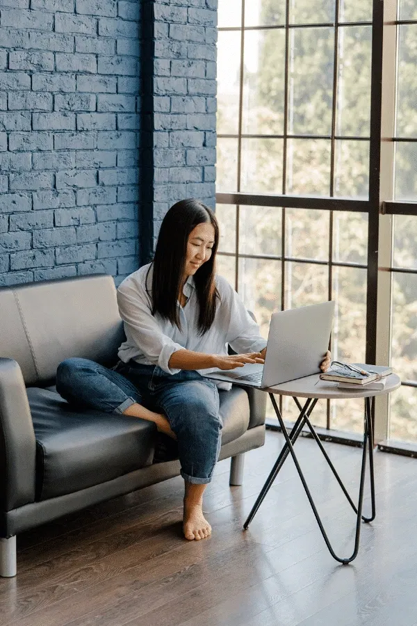 A woman sits barefoot on a gray sofa, working on a laptop placed on a small table by a large window in a modern room with blue brick walls.