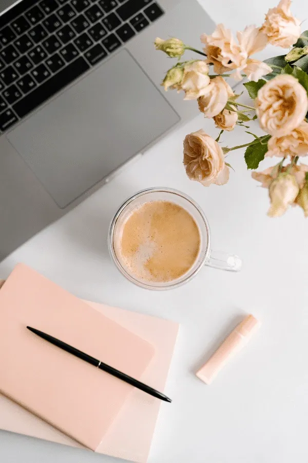 A laptop, a cappuccino in a glass mug, a closed notebook with a pen, a tube of lip balm, and a vase of pale peach roses on a white desk.