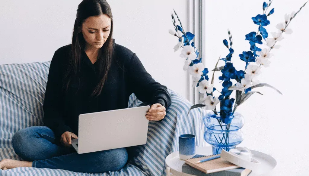 A woman sits on a striped couch using a laptop, with a table nearby holding a vase of blue and white flowers, a notebook, pen, and a blue cup.