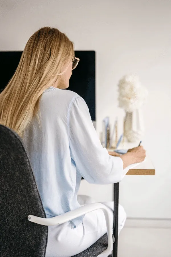 A woman with long blonde hair sits at a desk, writing on paper, facing away from the camera. A computer monitor and decorative items are on the desk.
