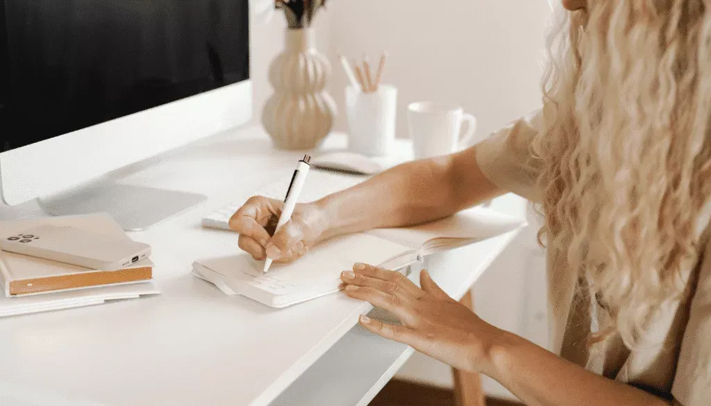 A person with long, curly hair writes in a notebook at a white desk with a computer, notebook, smartphone, and coffee mug.