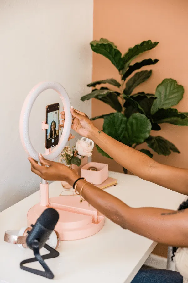 A person adjusts a smartphone on a ring light stand at a white desk with a microphone, flowers, and a plant in the background.