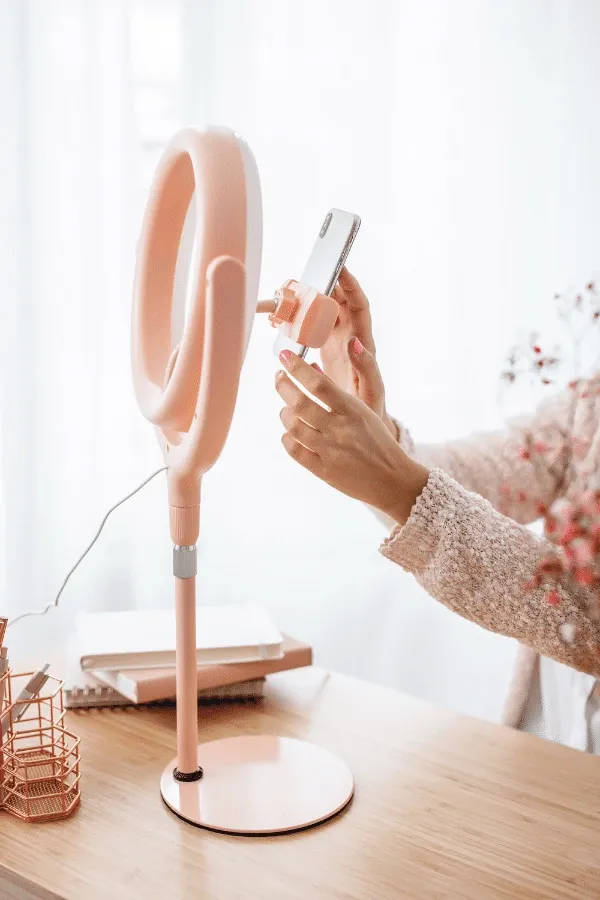 A person adjusts a smartphone on a ring light stand at a desk with notebooks and stationery in soft natural light.