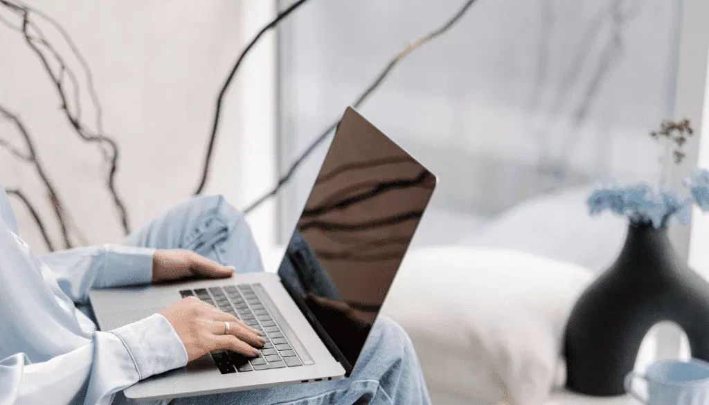 Person sitting with a laptop on their lap, typing, in a bright room with a black vase and blue flowers visible in the background.