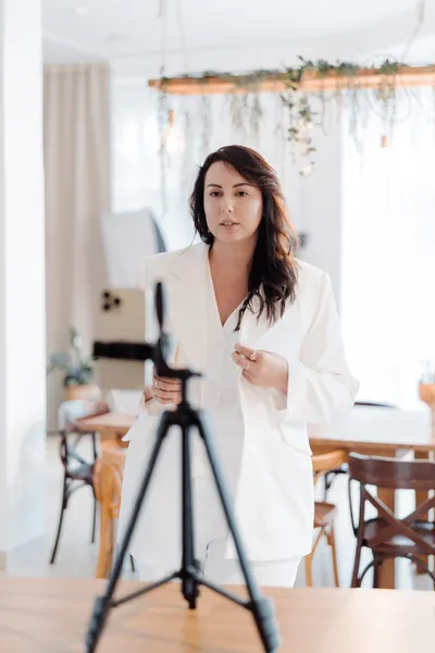 A woman in a white suit stands and speaks in front of a smartphone mounted on a tripod in a bright, modern room with wooden chairs and hanging plants.