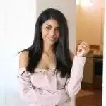 Woman with long dark hair in an off-shoulder light pink top stands indoors, smiling at the camera with her left hand slightly raised—perfect for a conscious copywriter’s profile picture.