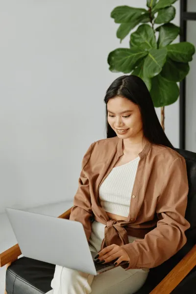 A woman sits on a black chair, using a laptop on her lap, with a large green plant in the background.
