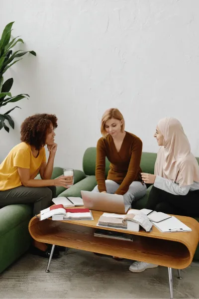 Three women sit around a coffee table with notebooks, a laptop, and drinks, engaged in conversation in a modern, bright room.